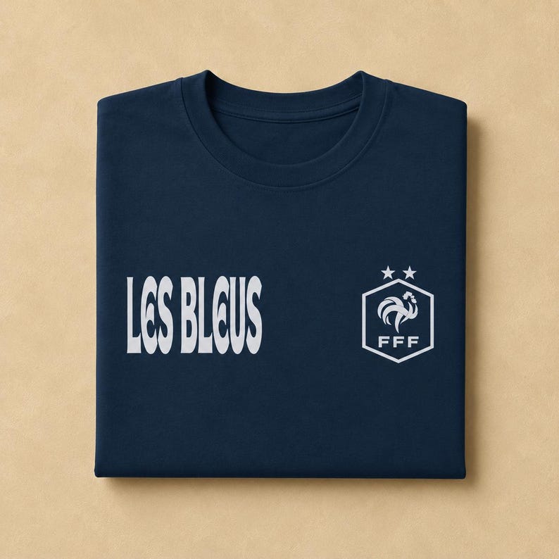 以下が含まれることがあります： 左側に白い文字で「LES BLEUS」、右側に白い六角形のロゴがプリントされたネイビーブルーのTシャツ。ロゴには、様式化された雄鶏、文字「FFF」、そして六角形の形状の上に配置された2つの星が描かれています。