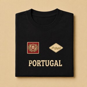 May include: A black crew neck t-shirt featuring a red square emblem with a gold crest and laurel wreath, a diamond-shaped patch with the word Portugal in script, and the word PORTUGAL printed in large gold capital letters below the graphics.