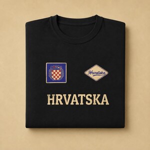 K&ouml;nnte beinhalten: Ein schwarzes T-Shirt mit Rundhalsausschnitt, das ein goldenes Wappen mit rot-wei&szlig;em Karomuster in einem blauen Quadrat, einen rautenf&ouml;rmigen Aufn&auml;her mit dem Text Hrvatska und das Wort HRVATSKA in gro&szlig;en goldenen Buchstaben darunter zeigt.