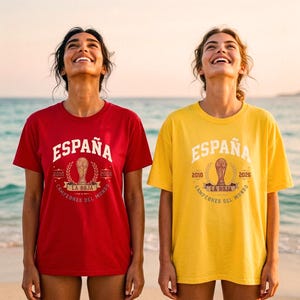 Può includere: Una t-shirt rossa e una t-shirt gialla, entrambe con una grafica bianca che riporta le scritte ESPAÑA, 2010, 2026, LA ROJA e CAMPEONES DEL MUNDO, centrate attorno all'illustrazione di un trofeo circondato da foglie di alloro.
