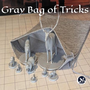 Könnte beinhalten: Ein grauer Kordelzugbeutel mit dem Text "Gray Bag of Tricks" enthält mehrere graue Tierfiguren, darunter ein Hirsch, Wölfe und kleinere Kreaturen. Die Tasche besteht aus einem strukturierten Stoff und ist offen, um den Inhalt zu zeigen.