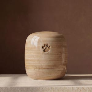 May include: A cylindrical ceramic urn with a glossy, light beige finish featuring horizontal wood-grain-like streaks and a small, debossed paw print centered on the front. The vessel has a smooth, rounded top and a flat base.