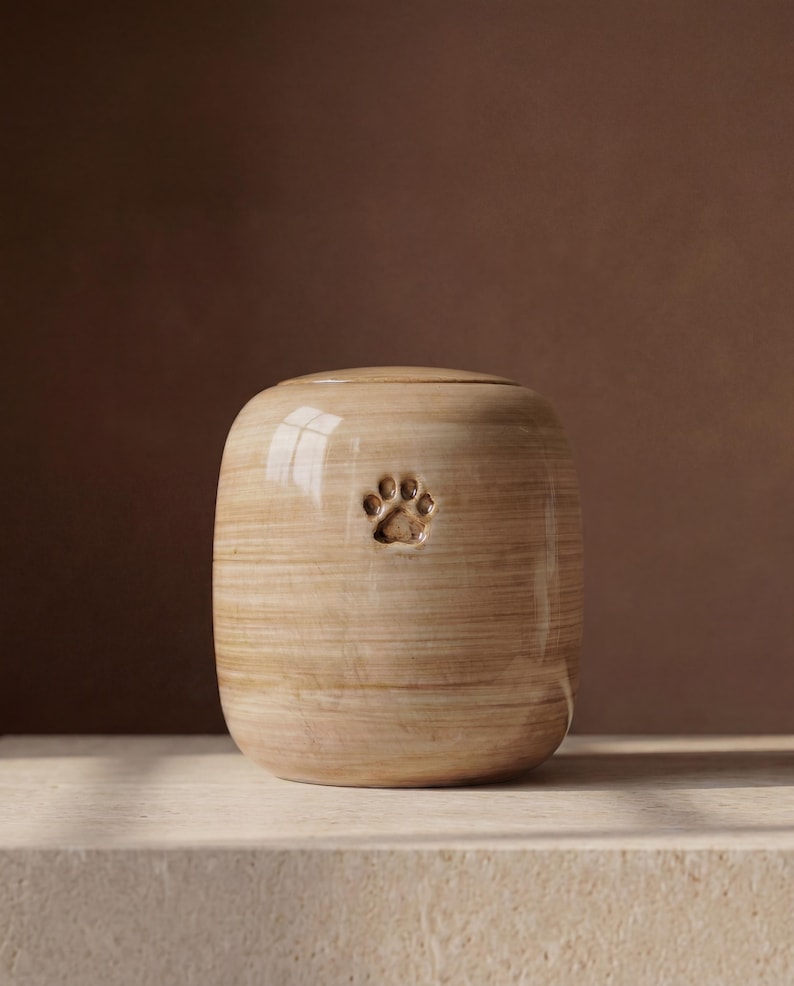 May include: A cylindrical ceramic urn with a glossy, light beige finish featuring horizontal wood-grain-like streaks and a small, debossed paw print centered on the front. The vessel has a smooth, rounded top and a flat base.
