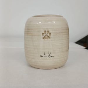 May include: A cylindrical ceramic urn with a light beige, horizontally brushed finish. The front features an embossed paw print above the engraved text Luli and Amor eterno in a dark script font, topped with a matching circular lid.