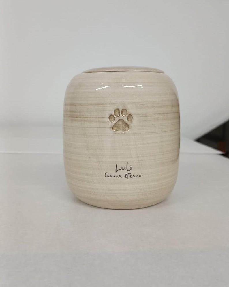May include: A cylindrical ceramic urn with a light beige, horizontally brushed finish. The front features an embossed paw print above the engraved text Luli and Amor eterno in a dark script font, topped with a matching circular lid.