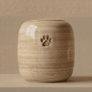May include: A cylindrical ceramic urn with a glossy, light beige finish featuring horizontal brushstroke textures. The front of the vessel displays a single, centered, debossed paw print indentation. The urn has a smooth, rounded top lid.