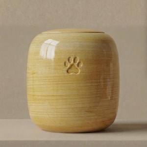 May include: A cylindrical ceramic urn with a glossy, light yellow finish and a subtle horizontal wood-grain texture. The front of the vessel features a small, debossed paw print centered near the top, and it includes a matching circular lid.