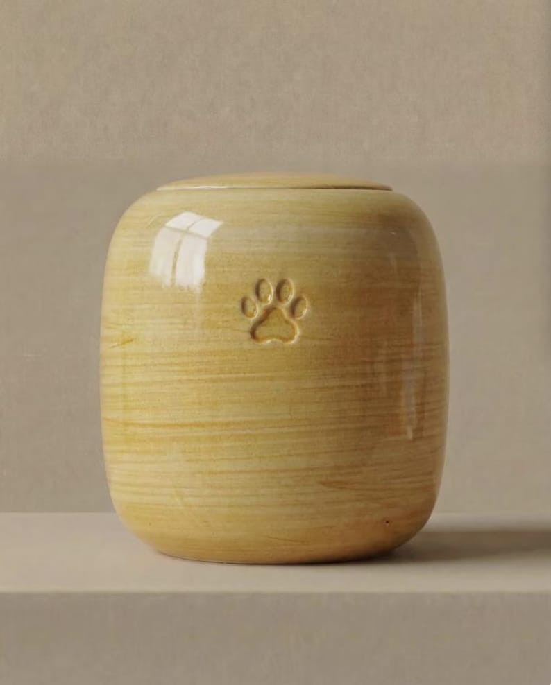 May include: A cylindrical ceramic urn with a glossy, light yellow finish and a subtle horizontal wood-grain texture. The front of the vessel features a small, debossed paw print centered near the top, and it includes a matching circular lid.