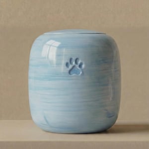 May include: A cylindrical ceramic urn with a light blue and white marbled finish. The exterior features a single embossed paw print centered on the front. The vessel has a smooth, rounded top lid and a consistent, hand-painted horizontal brushstroke pattern.