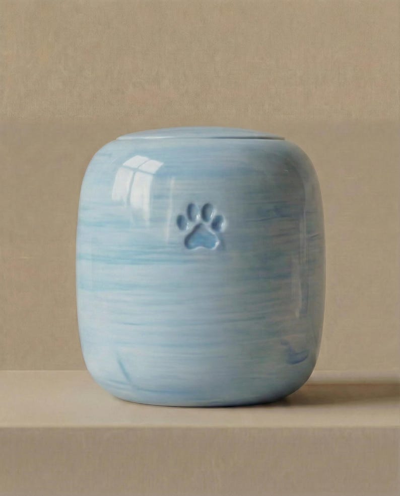 May include: A cylindrical ceramic urn with a light blue and white marbled finish. The exterior features a single embossed paw print centered on the front. The vessel has a smooth, rounded top lid and a consistent, hand-painted horizontal brushstroke pattern.
