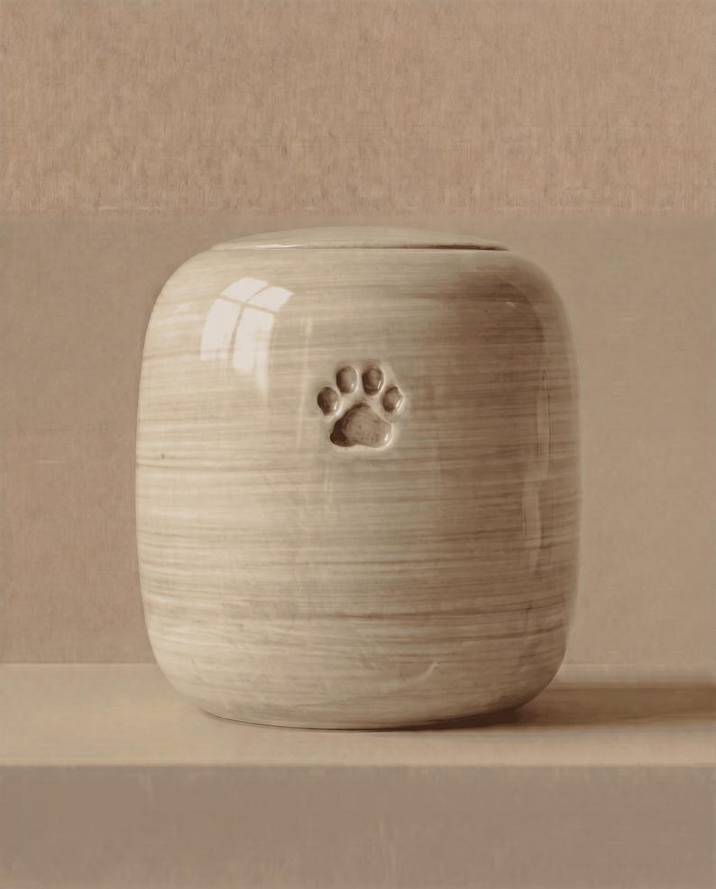 May include: A cylindrical ceramic urn with a smooth, glossy finish in a light beige tone featuring horizontal brushstroke textures and a single embossed paw print centered on the front surface, topped with a matching rounded lid.