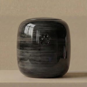 May include: A cylindrical ceramic urn with a glossy, dark gray and black marbled finish. The surface features a subtle, embossed paw print centered on the front. The vessel has a smooth, rounded top and a polished, reflective exterior texture.