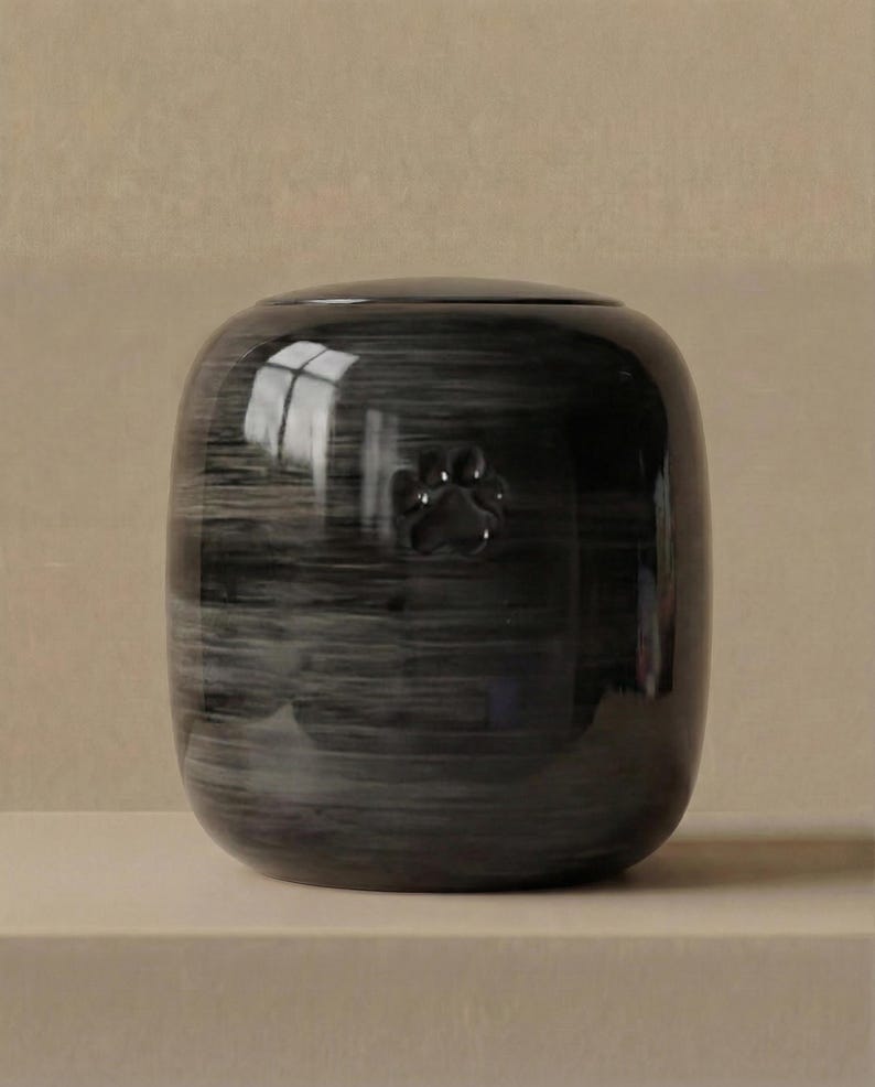 May include: A cylindrical ceramic urn with a glossy, dark gray and black marbled finish. The surface features a subtle, embossed paw print centered on the front. The vessel has a smooth, rounded top and a polished, reflective exterior texture.