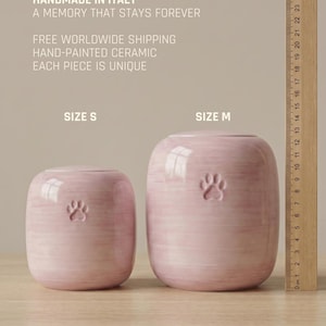 May include: Two light pink ceramic pet urns with a paw print design, labeled Size S and Size M. A wooden ruler shows the height of the larger urn is approximately 13 cm. The text reads: HANDMADE IN ITALY, A MEMORY THAT STAYS FOREVER, HAND-PAINTED CERAMIC.