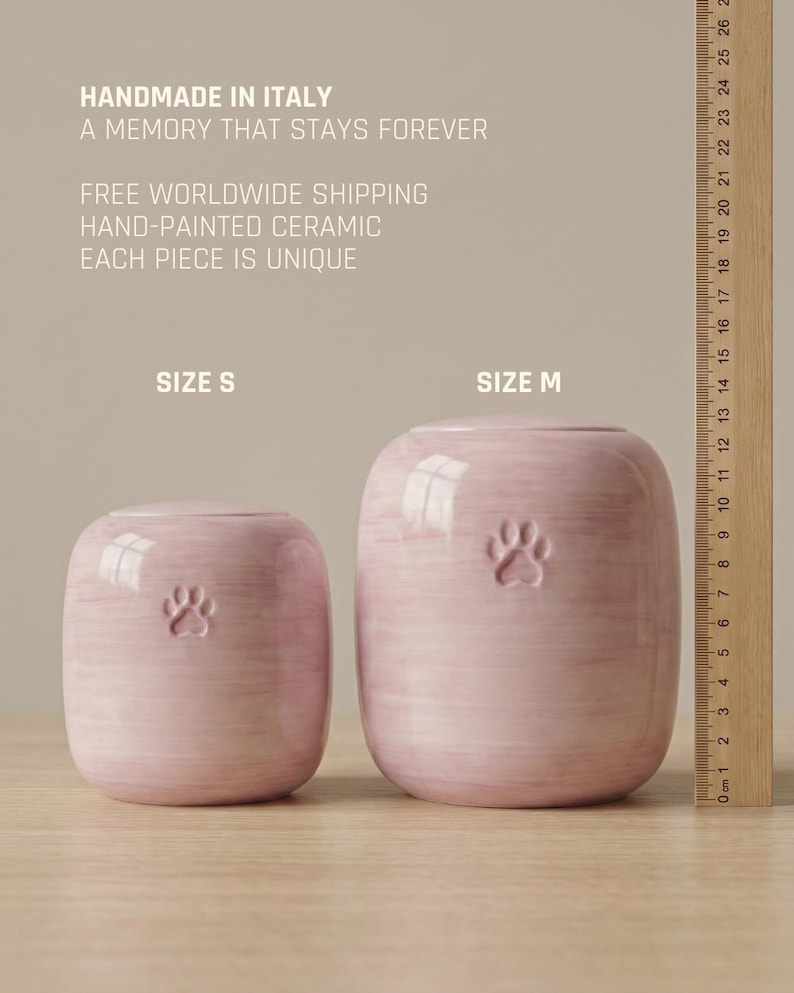 May include: Two light pink ceramic pet urns with a paw print design, labeled Size S and Size M. A wooden ruler shows the height of the larger urn is approximately 13 cm. The text reads: HANDMADE IN ITALY, A MEMORY THAT STAYS FOREVER, HAND-PAINTED CERAMIC.