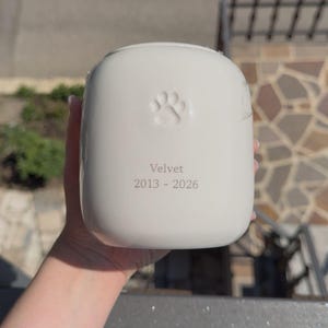 May include: A smooth, off-white ceramic urn with a rounded rectangular shape. The front features an embossed paw print centered above the engraved text "Velvet" and the years "2013 - 2026" in a simple, dark, sans-serif font.