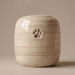 Op de afbeelding: Een cilindrische keramische urn met een glanzende, lichtbeige afwerking met horizontale penseelstreektexturen. De voorzijde van het vat toont een enkele, verdiepte pootafdruk in het midden, afgewerkt met een bijpassend rond deksel.