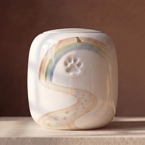 Può includere: Un'urna in ceramica bianca con un arcobaleno dipinto che sovrasta un sentiero di piccole impronte di zampe. Una piccola sagoma di cane è seduta sopra l'arcobaleno e un'impronta in rilievo è centrata sopra il sentiero sulla superficie liscia.