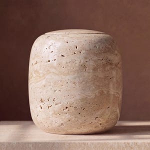 Op de afbeelding: Een cilindervormig vat gemaakt van beige travertijnsteen, met een poreus, getextureerd oppervlak met natuurlijke wervelpatronen en een bijpassend plat deksel. Het object heeft een glad, afgerond silhouet in een consistente lichtbruine kleur.