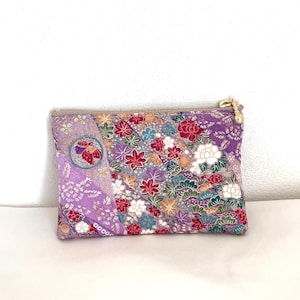 Puede incluir: Un estuche rectangular con cremallera que presenta un diseño de patchwork vibrante en tela morada, roja y verde. El tejido muestra patrones intrincados de hojas de arce, flores y motivos circulares con detalles dorados. Una pequeña cuenta dorada cuelga.