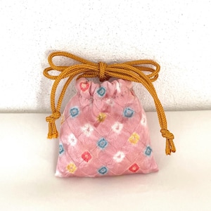 May include: A small pink drawstring pouch featuring a quilted diamond pattern and a repeating motif of tiny, multi-colored floral-like shapes in blue, yellow, white, and red. The bag is cinched at the top with a thick, mustard-yellow cord.