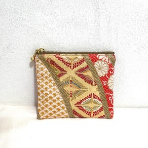 Puede incluir: Pequeño neceser cuadrado con cremallera y tirador dorados. La bolsa está hecha de tela con un diseño de patchwork. El diseño incluye secciones de tela dorada, roja y crema con varios patrones.