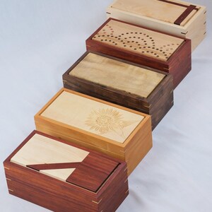 May include: A selection of wooden boxes with hinged lids. The boxes feature various designs, including inlaid patterns, a sunflower carving, and contrasting wood accents. The boxes are rectangular and appear to be handcrafted.