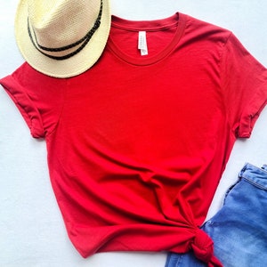 May include: A bright red t-shirt with rolled-up sleeves, paired with a straw hat and blue jeans. The hat has a black band. The t-shirt is knotted at the waist, creating a casual, stylish look. The items are arranged on a white surface.