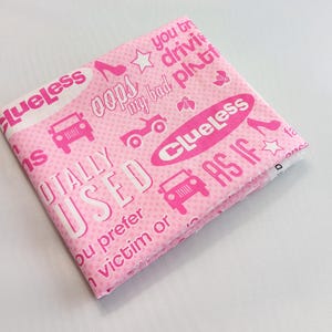 May include: Pink fabric with the word "Clueless" and other phrases and graphics in white and pink. The design includes cars, high heels, and stars. The fabric is folded and appears to be a handkerchief or similar item.