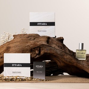 May include: A product display featuring fragrance oil and boxes. The boxes are white with a black band and the brand name "ITTARA". The fragrance oil bottle is clear with a silver cap. The boxes are labeled "Wood & Amber", "Musk & Amber", and "Silver Oud".