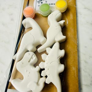 Peut inclure: Un kit de dinosaures à peindre soi-même. Le kit comprend quatre figurines de dinosaures en céramique blanche, trois petits pots de peinture orange, verte et jaune, et un petit pinceau. Le kit est emballé dans un sac en plastique transparent avec un dos en papier brun.