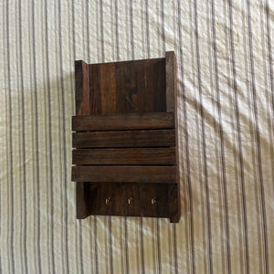 May include: A dark brown wooden wall organizer featuring a flat back panel, a horizontal storage slot with three wooden slats, and a lower section equipped with three small metal hooks for hanging items. The wood grain is visible throughout the piece.
