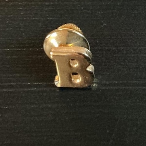 May include: A gold-toned metal lapel pin shaped like the capital letter B. The pin features a smooth, reflective surface and includes a circular clutch backing attached to the rear, designed to secure the letter to fabric or other materials.