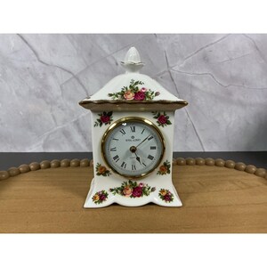 May include: A white porcelain mantel clock with a gold-trimmed roof and finial. The clock face features black Roman numerals and the text Royal Albert Quartz. The body is decorated with colorful pink, yellow, and red rose floral patterns.
