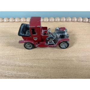 Pode incluir: Um carro de brinquedo vintage de metal fundido com acabamento em vermelho escuro, teto e assentos pretos. O veículo apresenta um capô dianteiro preto, rodas prateadas, um volante e um pequeno emblema em forma de escudo na porta lateral.