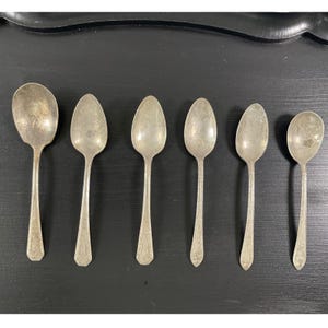 Peut inclure: Un ensemble de six cuillères en métal argenté disposées en rangée, de la plus grande à la plus petite. Chaque cuillère présente un manche simple et allongé avec un motif décoratif subtil à la base. Les surfaces métalliques sont ternies.
