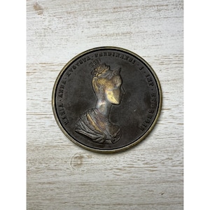 May include: A circular bronze-toned commemorative medal featuring a relief profile of a woman wearing a crown and necklace. The inscription reads MARIA ANNA AVGVSTA FERDINANDI I IMP ET REGIS. The medal has a dark patina with polished highlights.