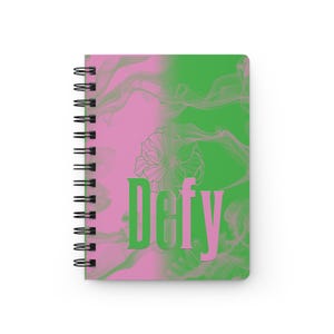 May include: Spiral-bound notebook with a pink and green abstract design. The cover features the word "Defy" in green and pink, with a floral outline. The notebook has a black spiral binding.