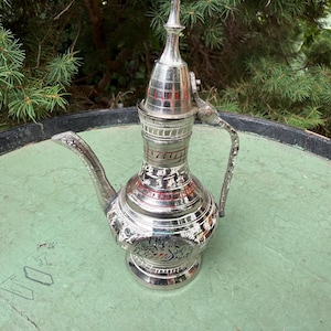 May include: A silver-colored metal ewer featuring an elongated spout, a curved handle, and a pointed lid. The body is decorated with intricate engraved patterns and red accents, showcasing a bird motif on the rounded base and a narrow neck.