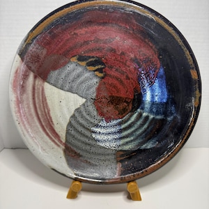 May include: A round ceramic plate featuring a multi-colored glaze with swirling patterns of deep red, dark blue, black, and speckled white. The surface has a textured, rippled appearance with a glossy finish, and the plate is supported by a small wooden stand.