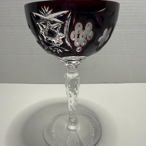 May include: A deep red and clear cut crystal wine glass featuring intricate starburst and grape cluster engravings on the bowl. The stem is faceted with a clear glass texture, leading down to a circular base with a radial pattern etched into the glass.