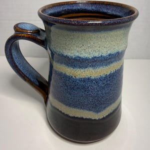 May include: A ceramic mug featuring a speckled glaze with horizontal bands of deep blue, light cream, and dark brown. The mug has a wide, flared rim and a sturdy, curved handle with a small, rounded thumb rest at the top of the handle attachment.
