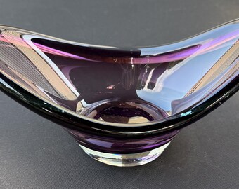 May include: A deep purple glass bowl with an elongated, curved, boat-like shape. The bowl features a thick, clear glass base that transitions into a translucent violet body with a smooth, polished rim and a reflective, glossy finish throughout.