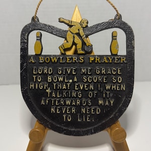 May include: A black and yellow cast metal wall plaque titled A BOWLERS PRAYER. It features a raised yellow figure of a bowler, two bowling pins, and the text: LORD GIVE ME GRACE TO BOWL A SCORE SO HIGH, THAT EVEN I, WHEN TALKING OF IT AFTERWARDS MAY NEVER NEED TO LIE.