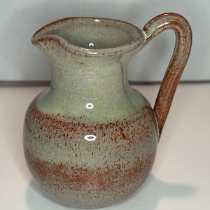 May include: A ceramic pitcher with a rounded body and a curved handle. The surface features a speckled glaze with a blend of light sage green and earthy reddish-brown tones. The pitcher has a wide opening with a small pouring spout at the top rim.
