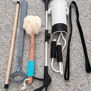 May include: A collection of daily living aids including a wooden dressing stick, a gray plastic shoehorn, a beige mesh bath sponge with a wooden handle, a black and silver reacher grabber tool, a white sock aid, and a black fabric leg lifter strap.