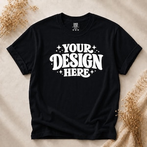 May include: A black short-sleeved t-shirt featuring the white text "YOUR DESIGN HERE" in a bold, rounded font, accented by small star and dot graphics. The shirt has a crew neckline and a visible interior label reading "COMFORT COLORS" and "M M M".
