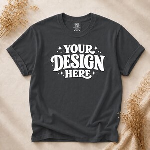 May include: A charcoal grey short-sleeve t-shirt featuring the white text "YOUR DESIGN HERE" in a bold, rounded font, accented by small star icons. The shirt has a crew neckline and a visible brand label on the inside collar marked with size M.