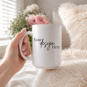 Puede incluir: Una taza de cerámica blanca con un asa grande, sostenida por una mano. La taza tiene el texto "Your design Here" en escritura negra. El fondo incluye una almohada esponjosa de color crema y un ramo de flores rosas y blancas.