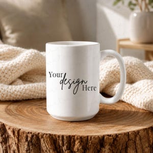 May include: A white ceramic mug with a large handle, resting on a wooden surface. The mug features the text "Your design Here" in black script. The background includes a cream-colored blanket and a blurred backdrop.